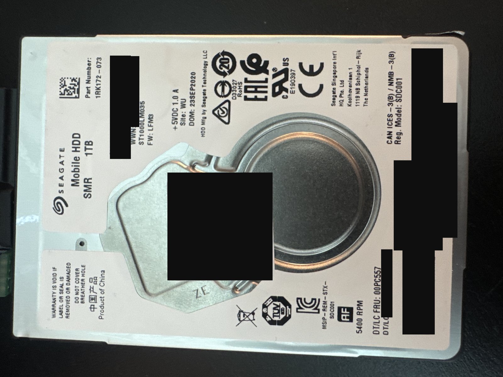 Seagate ST1000LM035 1 TB SMR laptop hard drive label with serial number, WWN, barcodes, PSID and data-matrix redacted, and a prior lab's intake sticker covered