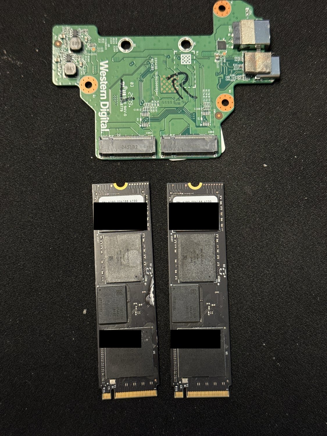 Two WD_BLACK SN850X M.2 NVMe SSDs and the two-bay USB enclosure bridge board they were striped across, with each SSD's serial number, PSID and data-matrix codes redacted